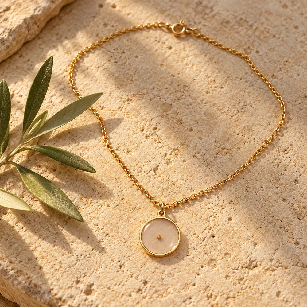 Gold-tone round mustard seed pendant on a fine chain on a pale limestone background, with an olive sprig for scale — true size: the round pendant is about 18 mm across, with one tiny 2 mm mustard seed in the centre, on a fine cable chain about 1 mm thick