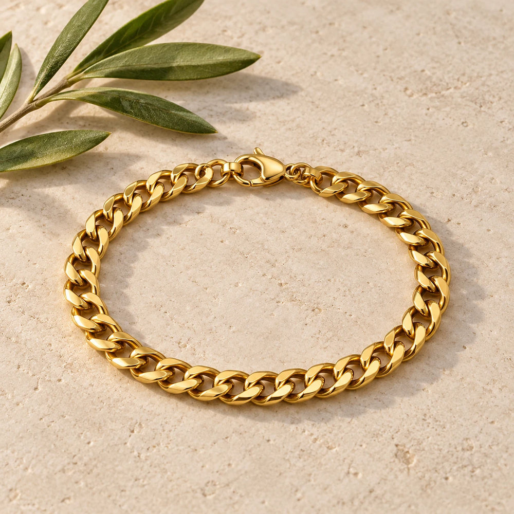 Gold-tone 6 mm cuban chain bracelet on a pale limestone background, with an olive sprig for scale — true size: the flat curb bracelet is 6 mm wide with 1.6 mm wire, 21 cm long