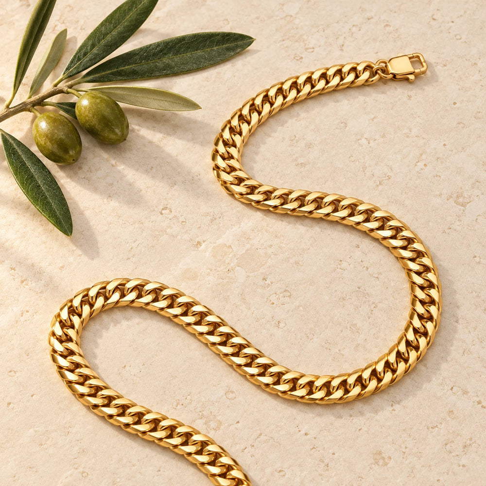 Gold-tone 6 mm cuban chain necklace on a pale limestone background, with an olive sprig for scale — true size: the dense cuban chain is 6 mm wide
