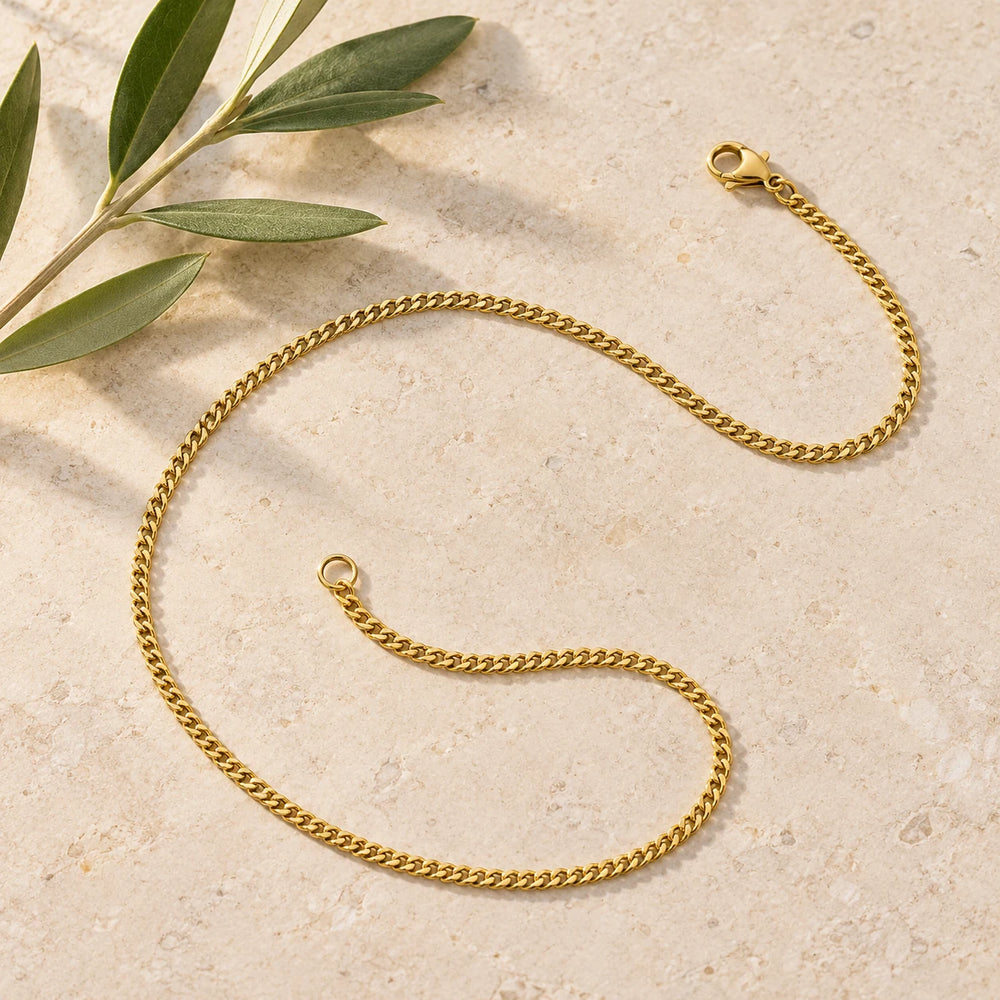 Gold-tone 2 mm cuban chain on a pale limestone background, with an olive sprig for scale — true size: the curb chain is 2 mm wide