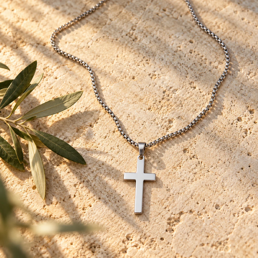 Silver-tone polished cross pendant on a box chain on a pale limestone background, with an olive sprig for scale — true size: the plain cross is 43 mm tall and 24 mm wide (about 50 mm with the bail), on a box chain about 2.5 mm wide