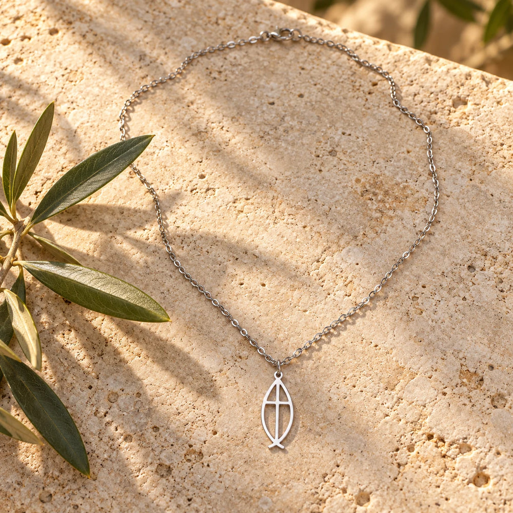 Silver-tone ichthys fish pendant with a cross inside on a fine chain on a pale limestone background, with an olive sprig for scale — true size: the open fish pendant is about 23 mm long and 10 mm wide, slim and flat, on a fine cable chain about 1 mm thick