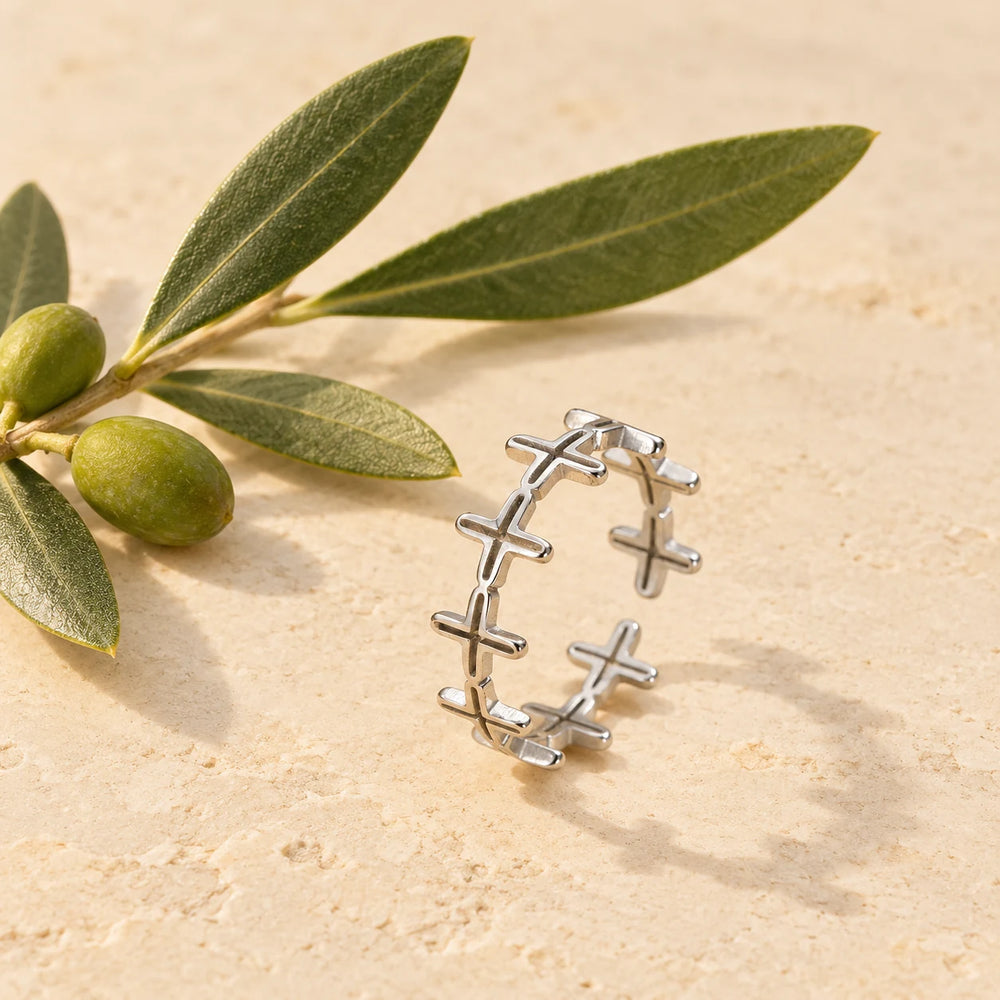 Silver-tone band of small crosses ring on a pale limestone background, with an olive sprig for scale — true size: the band is a single row of tiny crosses 6 mm tall, the ring about 20 mm across