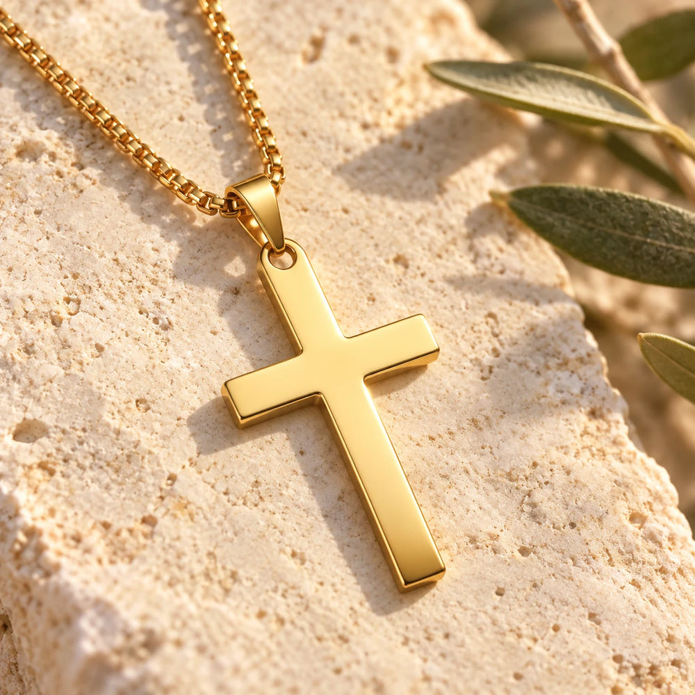 Close-up of the gold-tone polished cross pendant on a box chain on limestone beside olive leaves