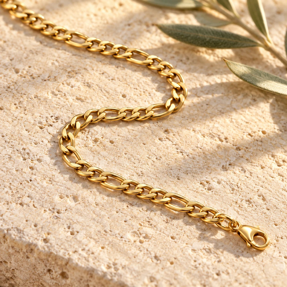 Close-up of the gold-tone 3 mm figaro chain necklace on limestone beside olive leaves