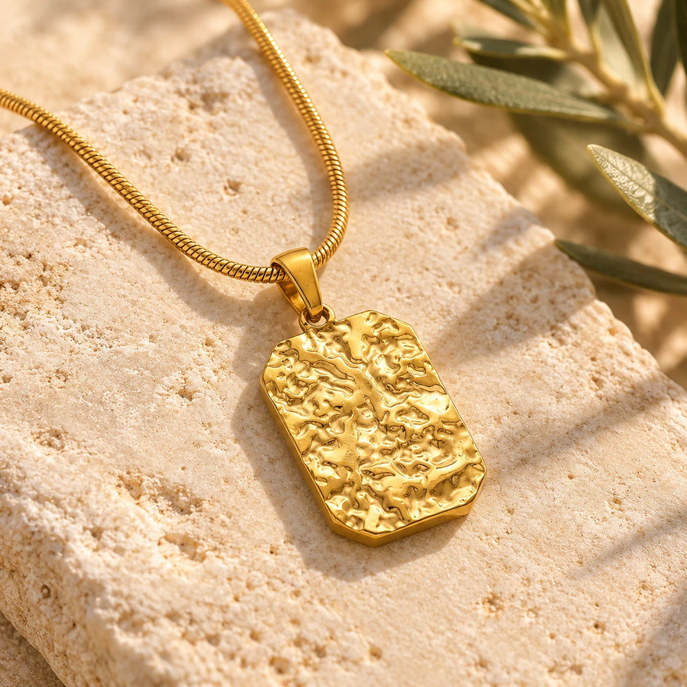 Close-up of the gold-tone stone-relief tag pendant on limestone beside olive leaves