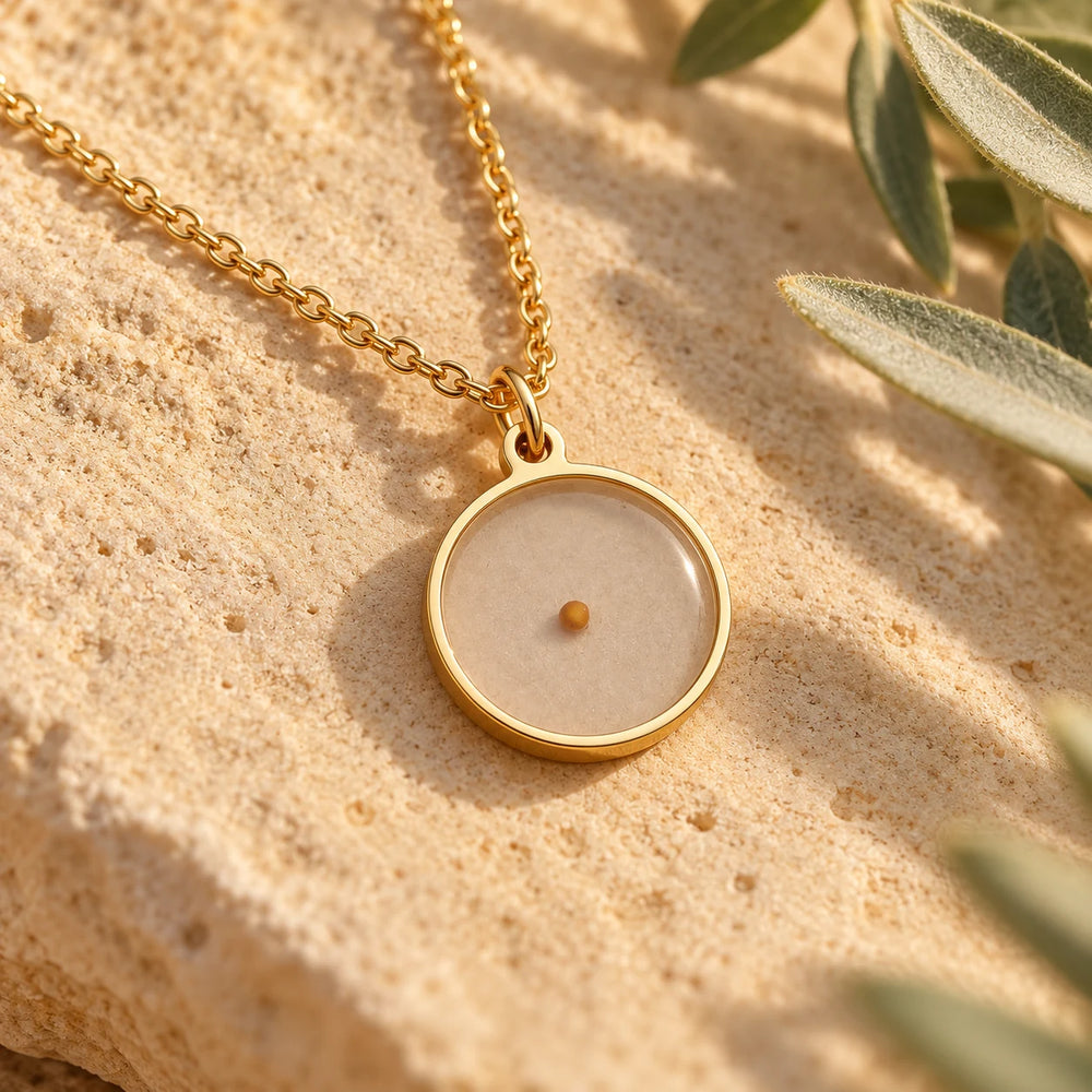 Close-up of the gold-tone mustard seed pendant on limestone beside olive leaves, the single seed visible under the clear dome