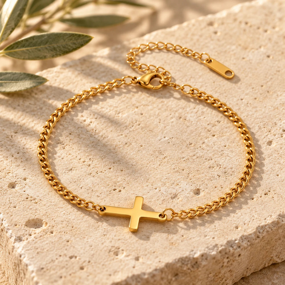 Close-up of the gold-tone curb chain bracelet with a sideways cross on limestone beside olive leaves