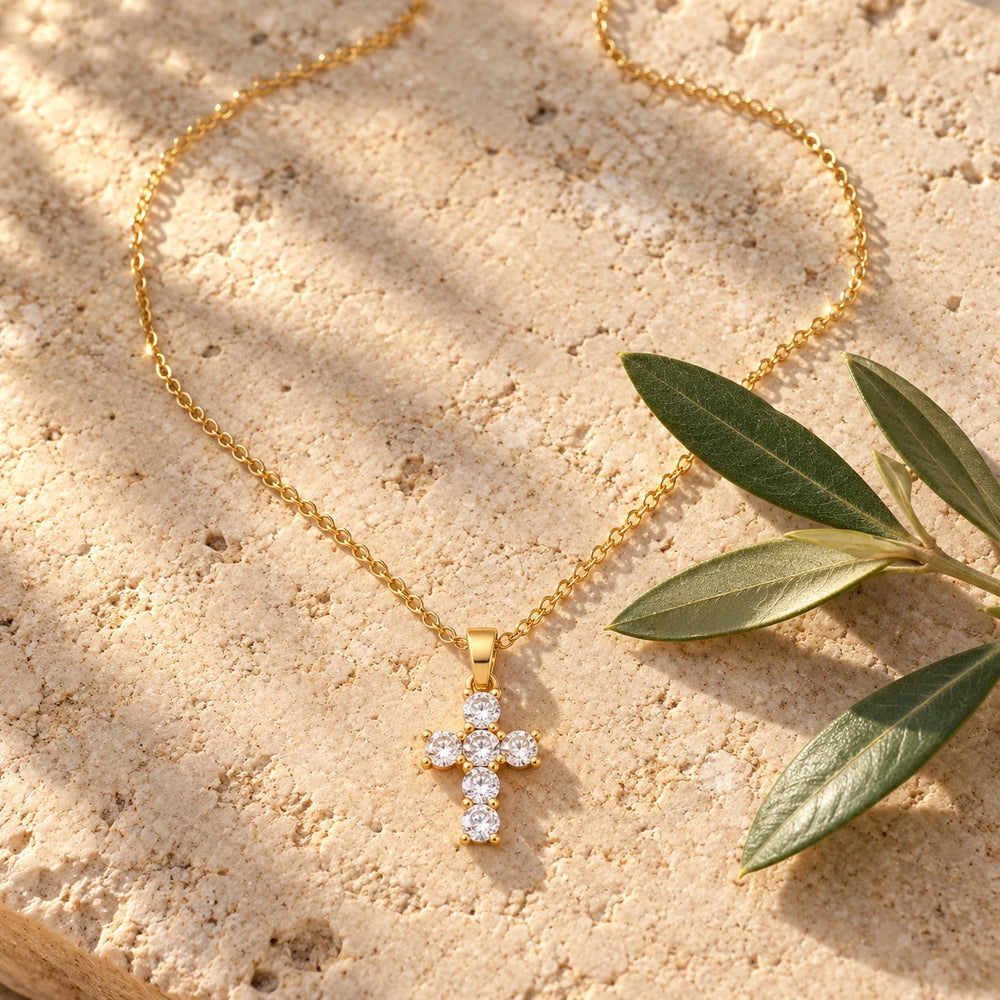 Gold-tone cross with six clear stones on a fine chain on a pale limestone background, with an olive sprig for scale — true size: the stone cross is small: 21 mm tall including its bail and 12 mm wide, each stone about 3.5 mm, on a fine cable chain about 1 mm thick