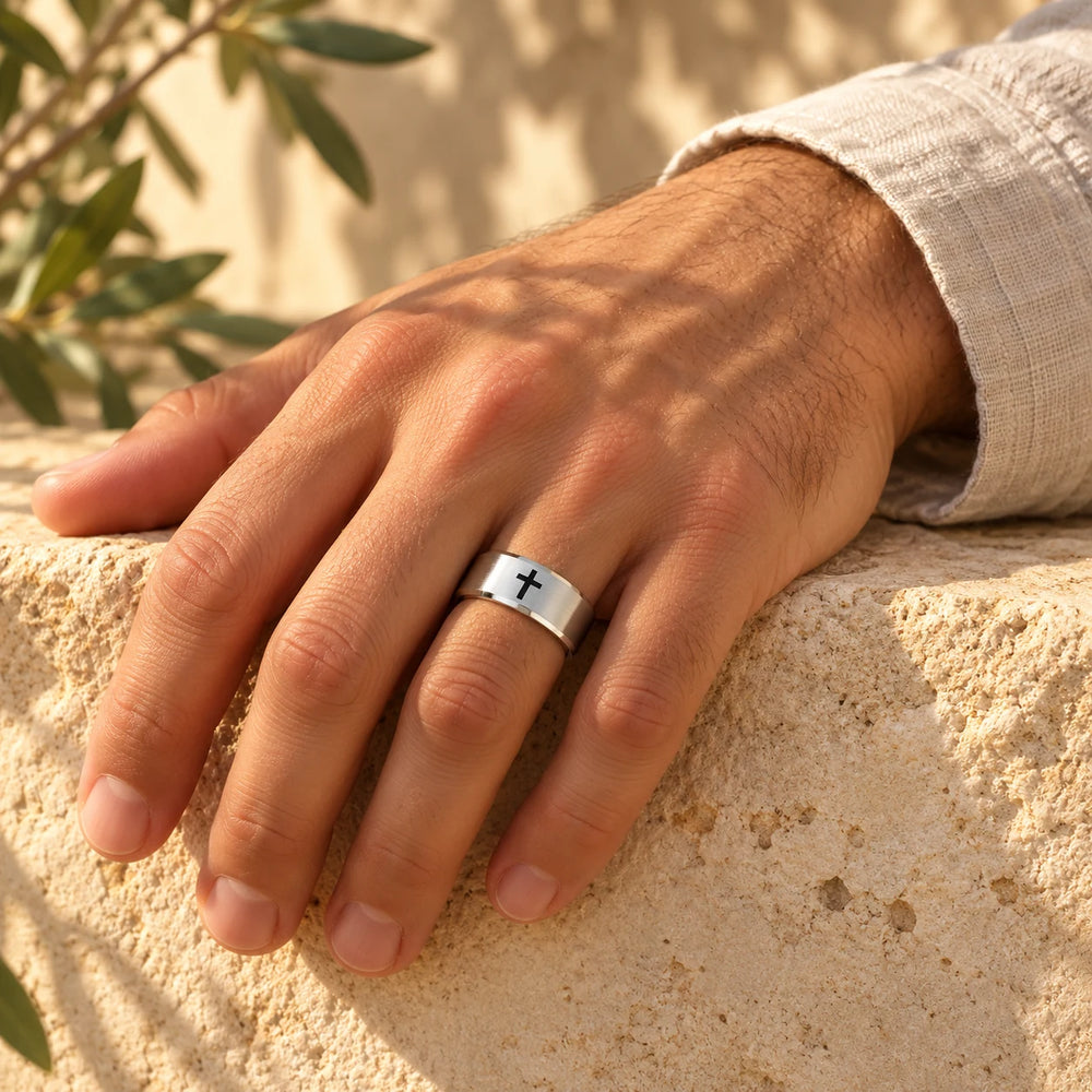 Silver-tone cross band ring on a man's hand resting on a limestone wall — true size: the band is 8 mm wide with an outer diameter of about 22–24 mm; the black cross on top is about 3–4 mm