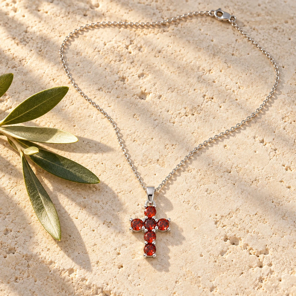 Silver-tone cross with six red stones on a fine chain on a pale limestone background, with an olive sprig for scale — true size: the stone cross is small: 21 mm tall including its bail and 12 mm wide, each stone about 3.5 mm, on a fine cable chain about 1 mm thick