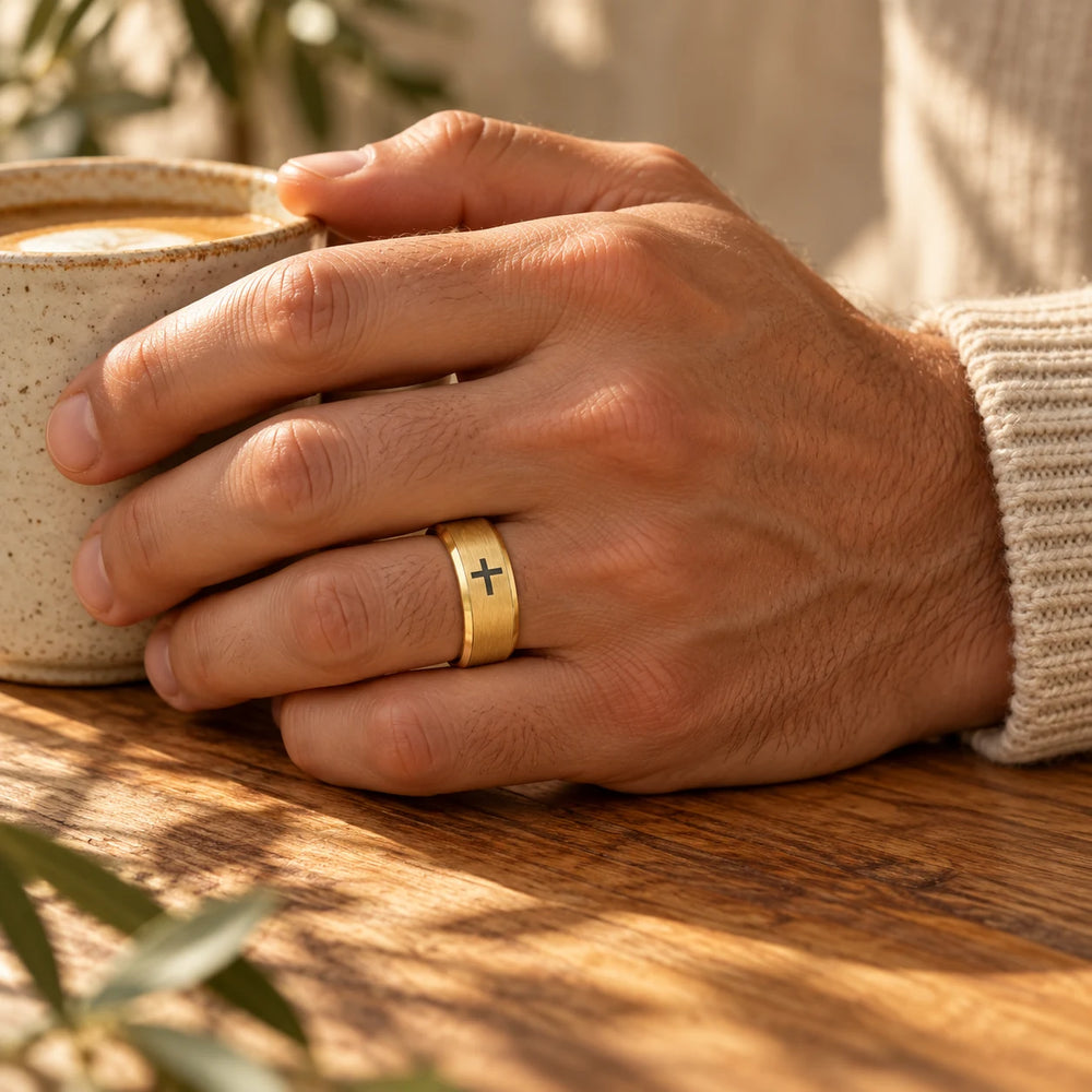 Gold-tone cross band ring on a man's hand holding a coffee cup — true size: the band is 8 mm wide with an outer diameter of about 22–24 mm; the black cross on top is about 3–4 mm