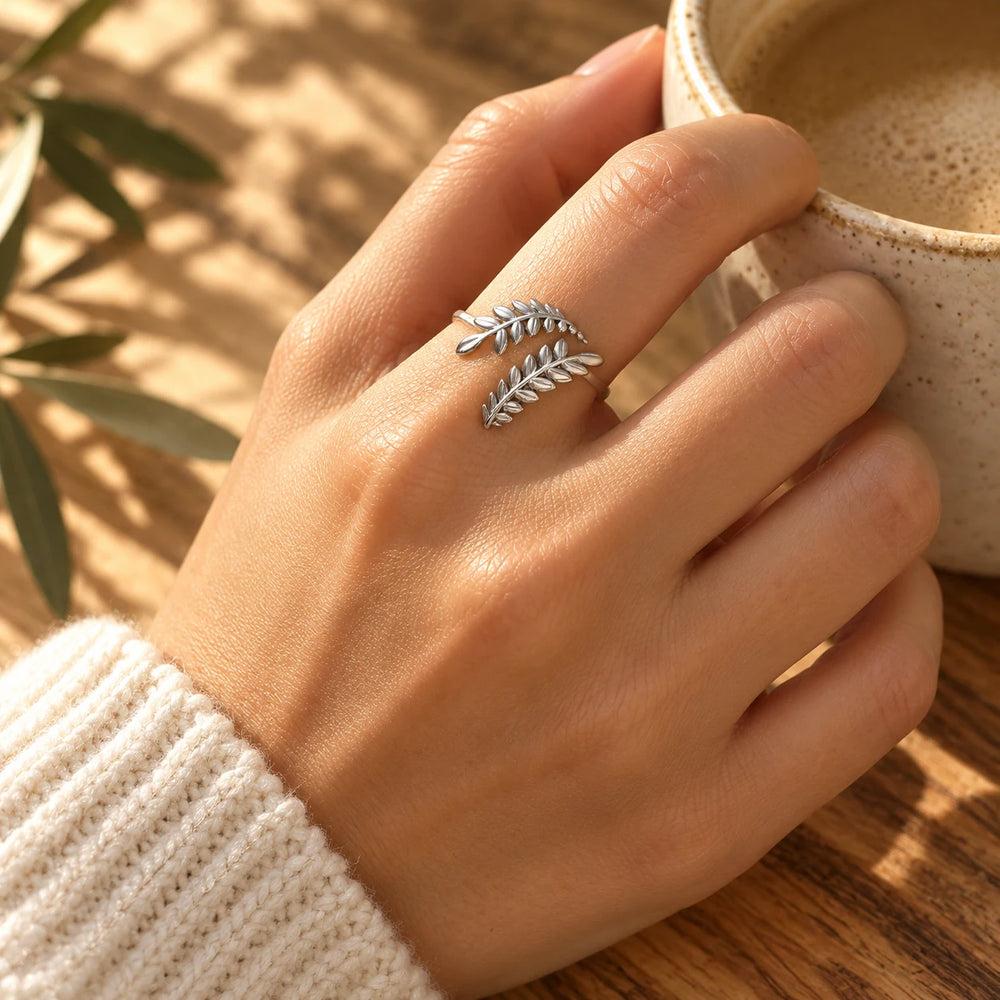Silver-tone olive branch ring on a woman's hand holding a ceramic cup — true size: the shank is about 1 mm thick and each olive sprig is about 5–6 mm wide; the ring is about 19 mm across