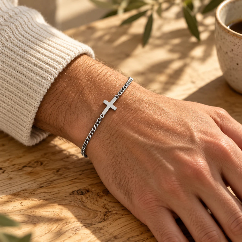 Silver-tone curb chain bracelet with a sideways cross on a man's wrist on an olive-wood table — true size: the sideways cross is 20 × 12 mm on a curb chain about 2.5 mm wide, 17 cm + 5 cm extender