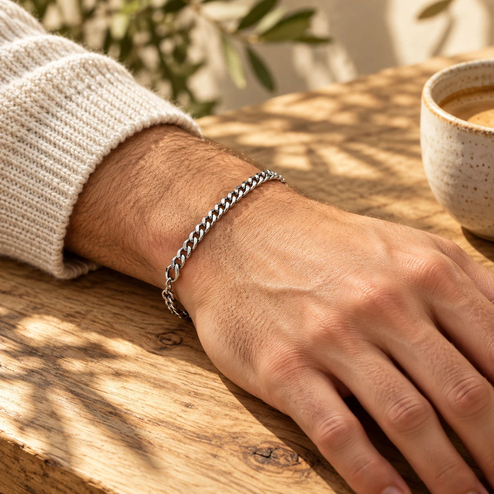 Silver-tone 4 mm cuban chain bracelet on a man's wrist on an olive-wood table — true size: the flat curb bracelet is 4 mm wide with 1 mm wire, 17 cm + 5 cm extender