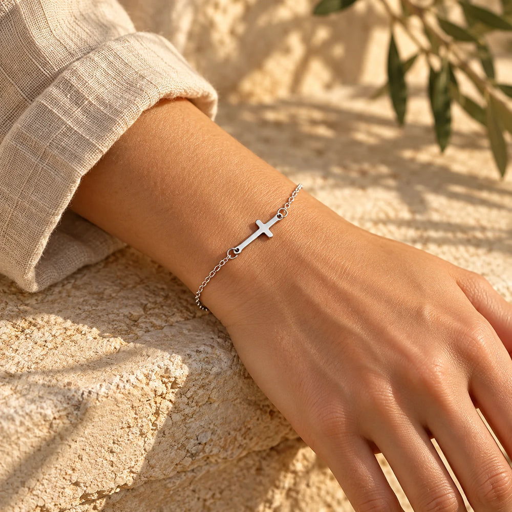 Silver-tone fine cross bracelet worn on a woman's wrist — true size: the slim sideways cross is about 21 mm long and 7 mm tall, on a fine cable chain about 1.5 mm wide