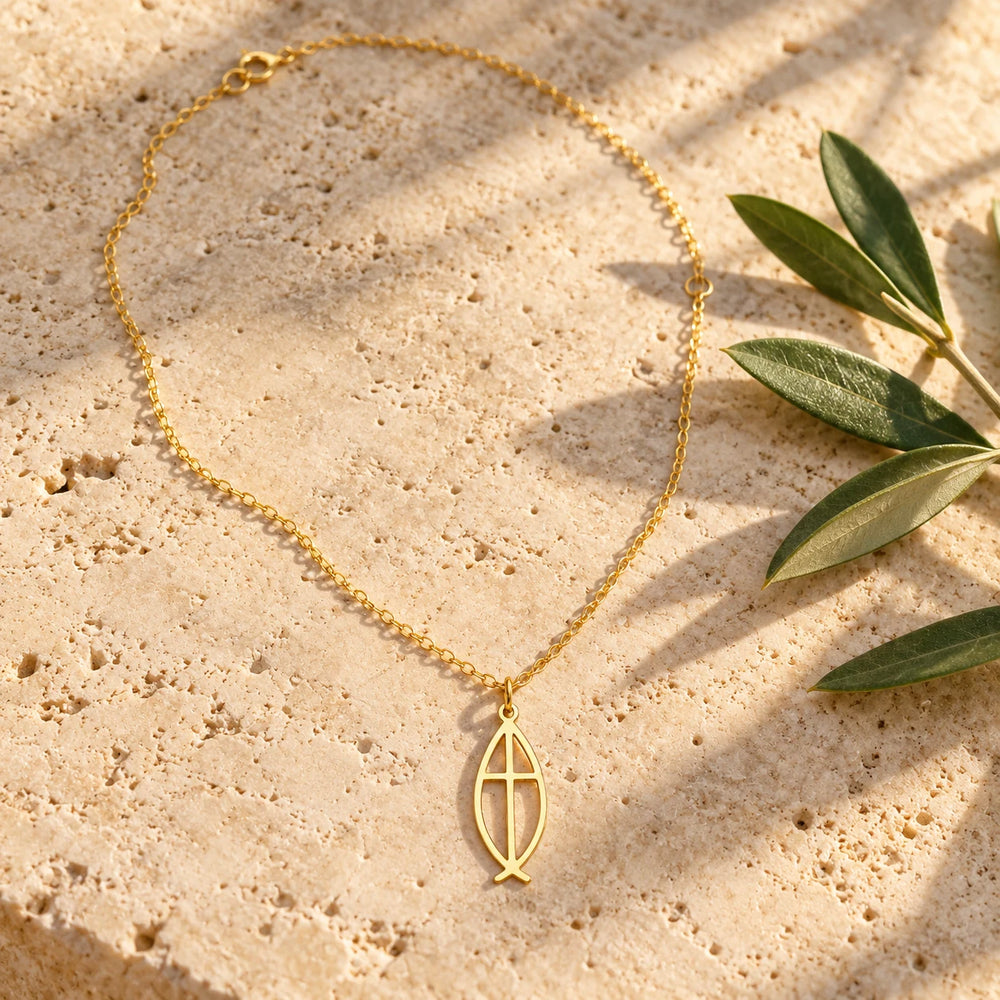Gold-tone ichthys fish pendant with a cross inside on a fine chain on a pale limestone background, with an olive sprig for scale — true size: the open fish pendant is about 23 mm long and 10 mm wide, slim and flat, on a fine cable chain about 1 mm thick