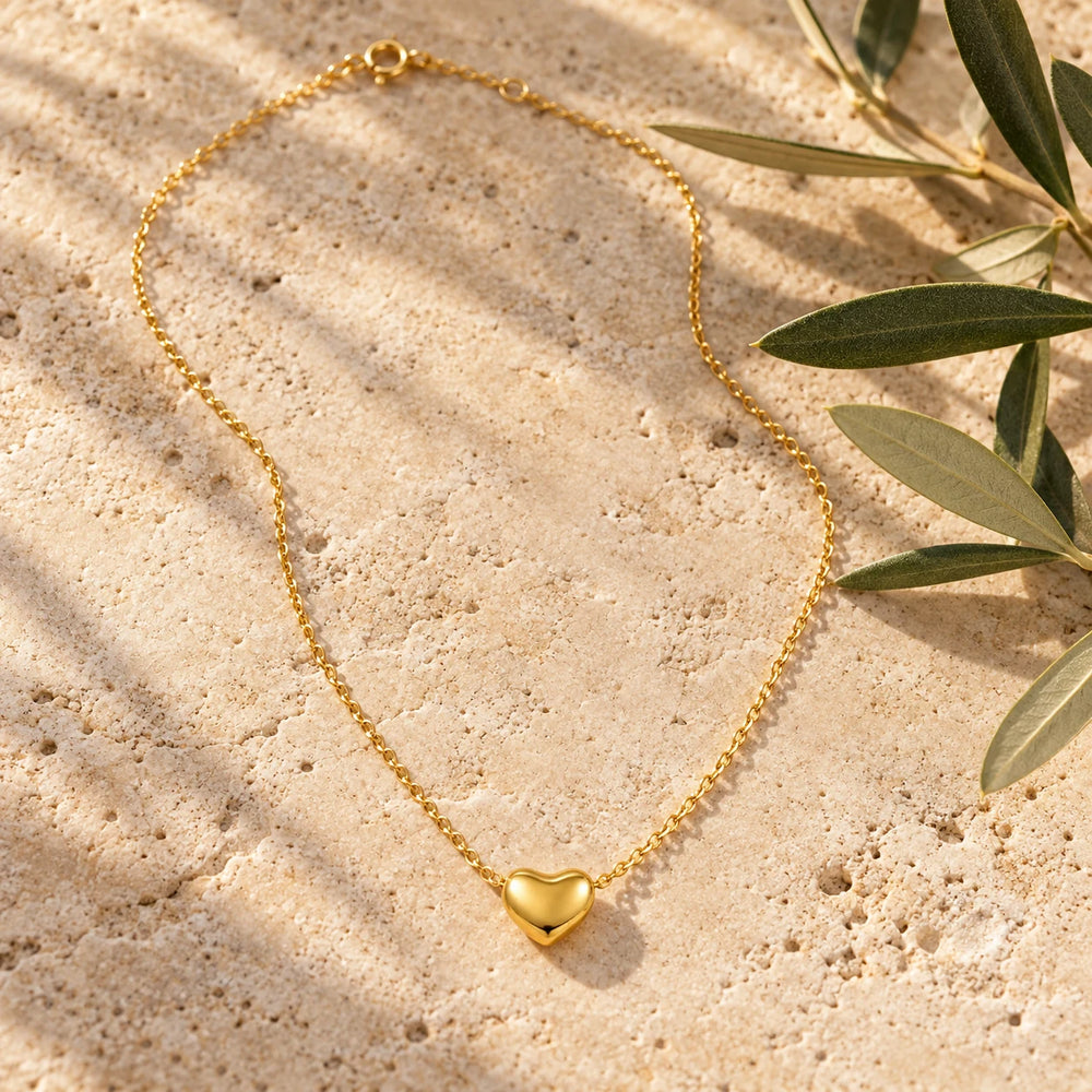Gold-tone small puffed heart on a fine chain on a pale limestone background, with an olive sprig for scale — true size: the puffed heart is about 10 mm wide, threaded on a fine cable chain about 1 mm thick
