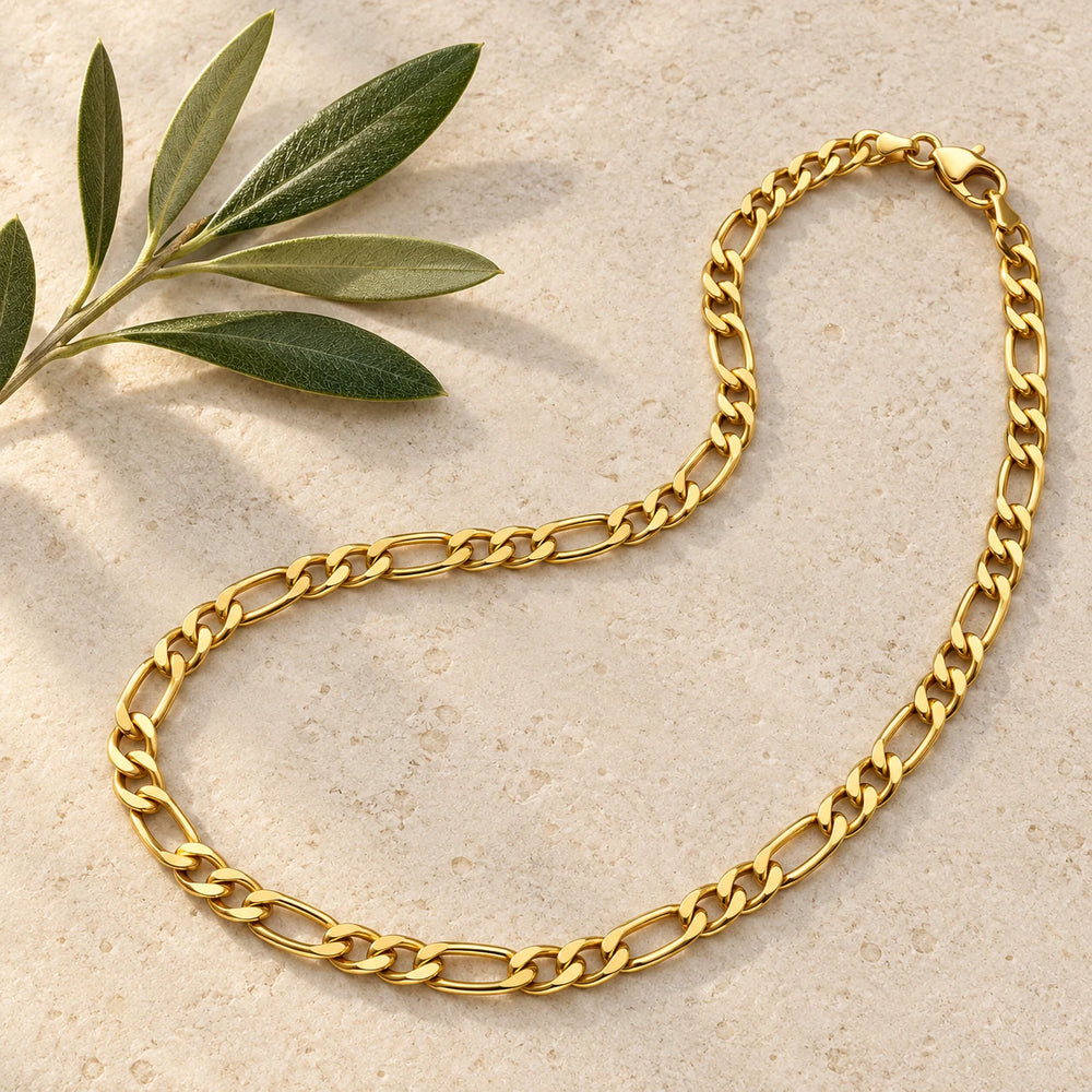 Gold-tone 5 mm figaro chain necklace on a pale limestone background, with an olive sprig for scale — true size: the figaro chain is 5 mm wide with about 1.4 mm wire