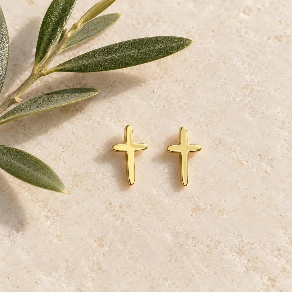 Pair of gold-tone small cross stud earrings on a pale limestone background, with an olive sprig for scale — true size: each cross stud is 14 mm tall and 8 mm wide
