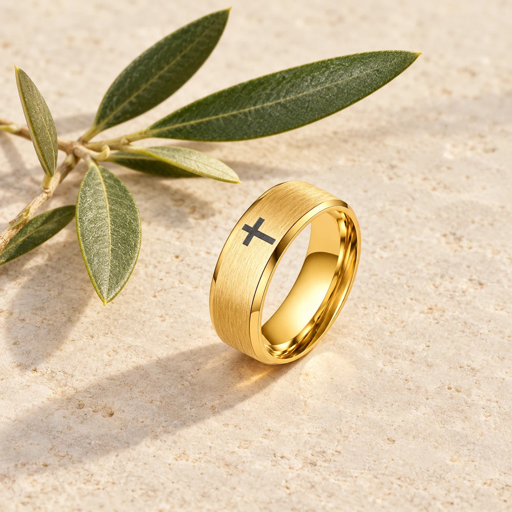 Gold-tone brushed band ring with a small black cross on a pale limestone background, with an olive sprig for scale — true size: the band is 8 mm wide with an outer diameter of about 22–24 mm; the black cross on top is about 3–4 mm