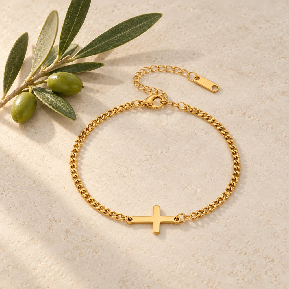 Gold-tone curb chain bracelet with a sideways cross on a pale limestone background, with an olive sprig for scale — true size: the sideways cross is 20 × 12 mm on a curb chain about 2.5 mm wide, 17 cm + 5 cm extender