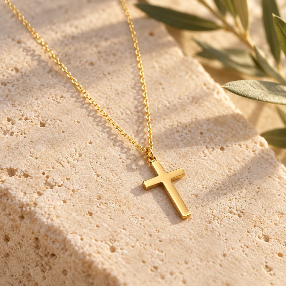 Close-up of the gold-tone dainty plain cross necklace on a fine chain on limestone beside olive leaves