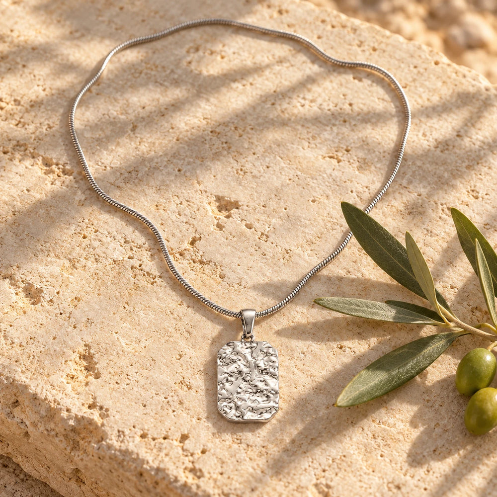 Silver-tone textured stone-relief tag pendant on a snake chain on a pale limestone background, with an olive sprig for scale — true size: the square textured tag is 22 mm tall and 16 mm wide (about 28 mm with the bail), on a fine chain about 1.5 mm thick