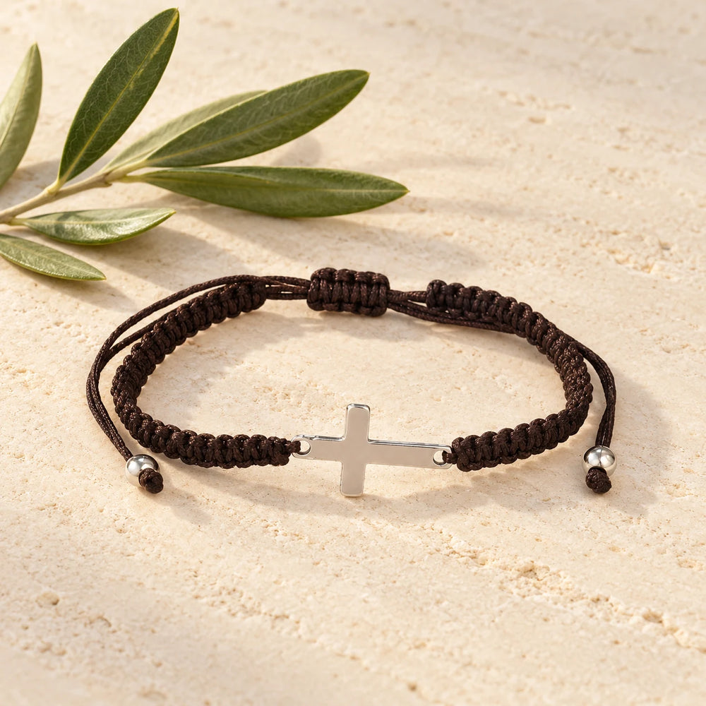 Dark brown braided cord bracelet with a small steel cross on a pale limestone background, with an olive sprig for scale — true size: the braided cord is about 5 mm thick and the sideways steel cross about 20 × 11 mm