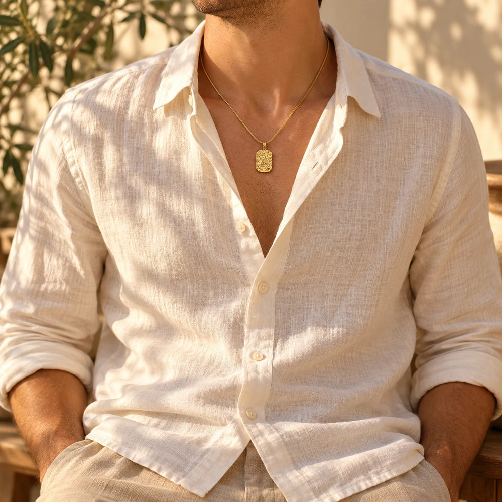 Gold-tone stone-relief tag pendant worn with an open linen shirt — true size: the square textured tag is 22 mm tall and 16 mm wide (about 28 mm with the bail), on a fine chain about 1.5 mm thick
