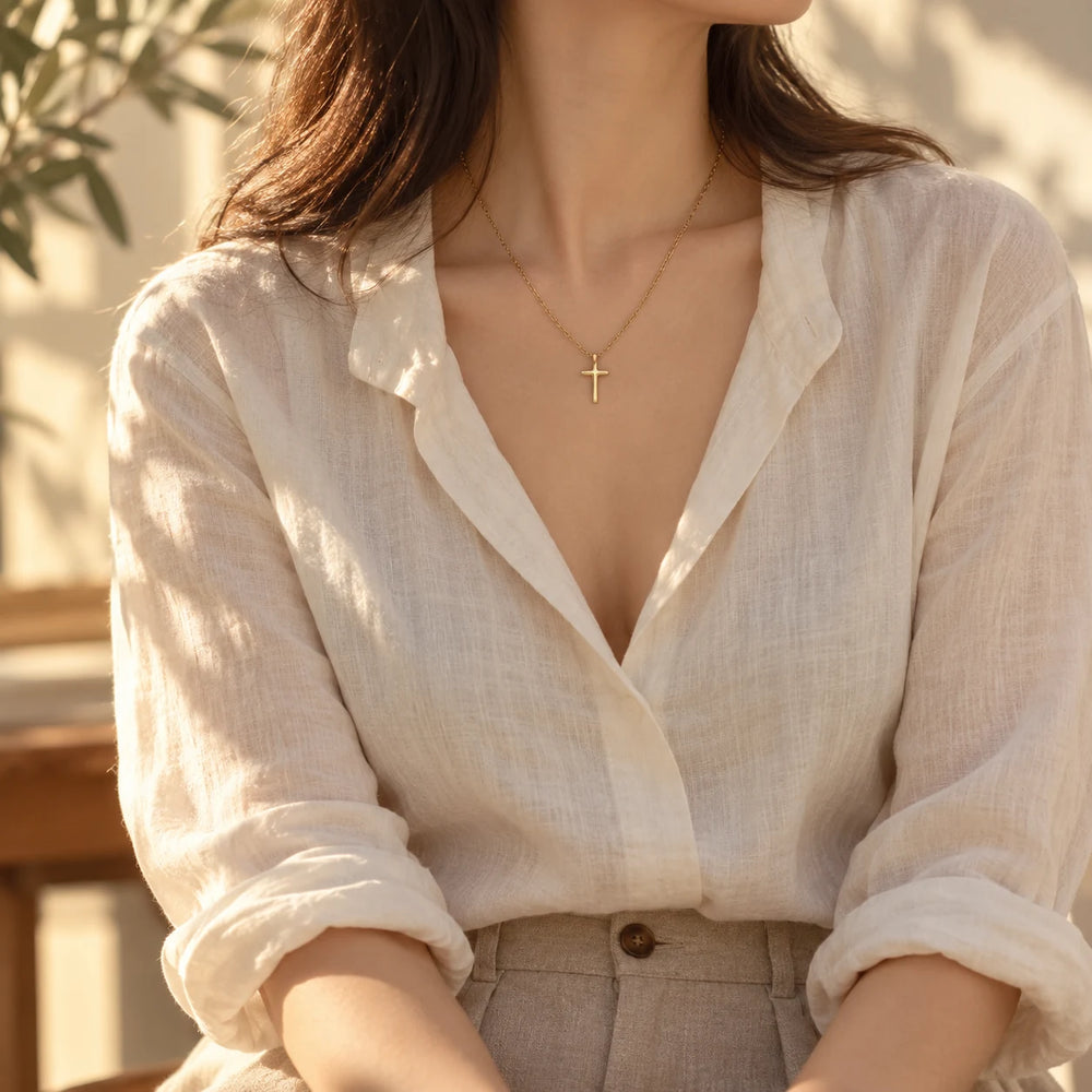 Gold-tone tiny cross necklace worn at the collarbone with a white cotton blouse — true size: the tiny cross is 14 mm tall and 8 mm wide, about the size of a fingernail, on a fine cable chain about 1 mm thick