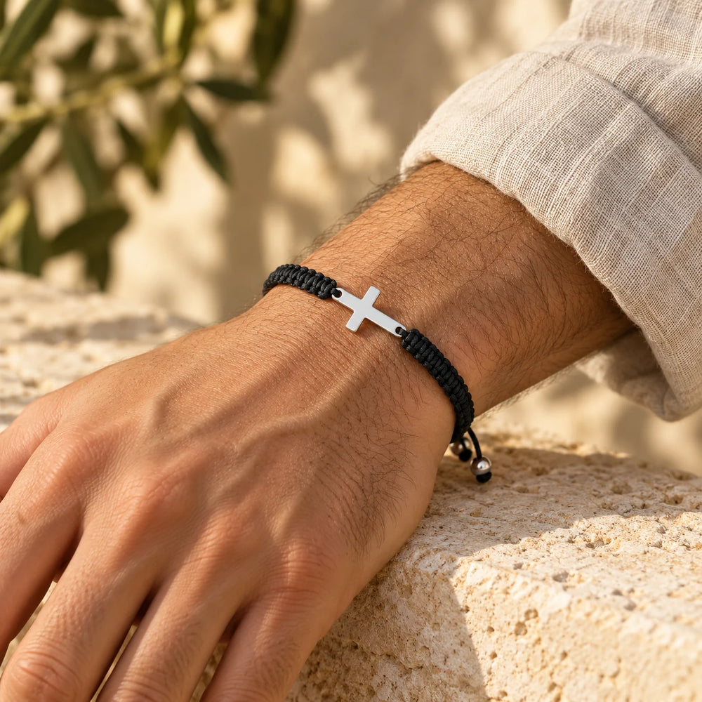Black cord cross bracelet on a man's wrist resting on a limestone wall — true size: the braided cord is about 5 mm thick and the sideways steel cross about 20 × 11 mm