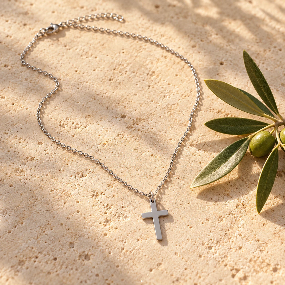 Silver-tone dainty plain cross on a fine chain on a pale limestone background, with an olive sprig for scale — true size: the cross is about 20 mm tall and 12–14 mm wide, on a fine chain about 1.3–2 mm wide