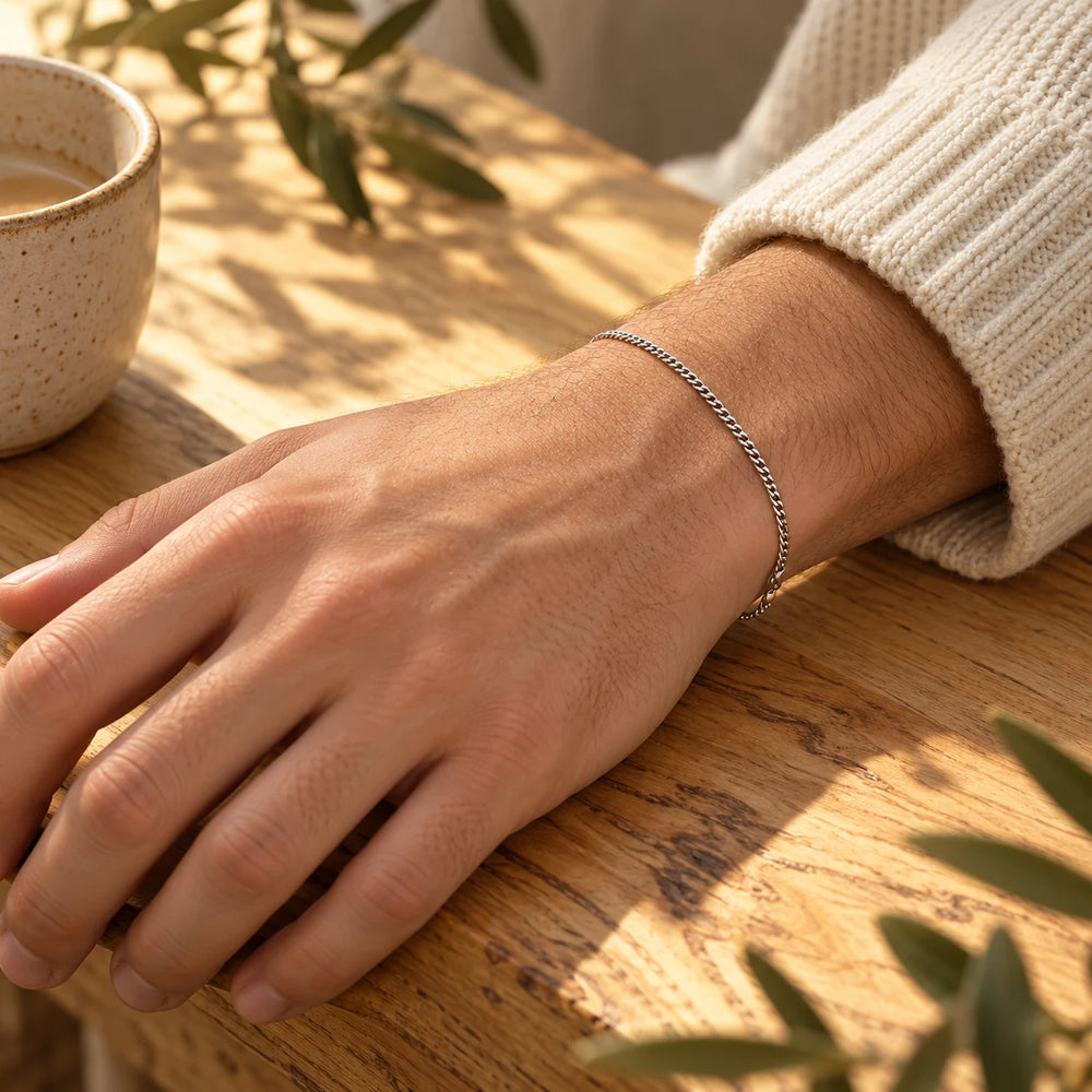Silver-tone 2.5 mm cuban chain bracelet on a man's wrist on an olive-wood table — true size: the curb bracelet is 2 mm wide, 20 cm long