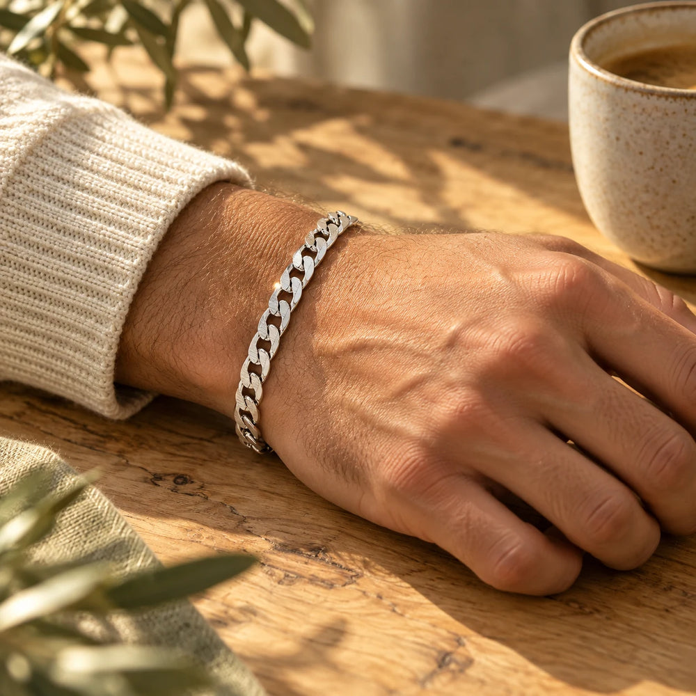Silver-tone 6 mm cuban chain bracelet on a man's wrist on an olive-wood table — true size: the flat curb bracelet is 6 mm wide with 1.6 mm wire, 21 cm long