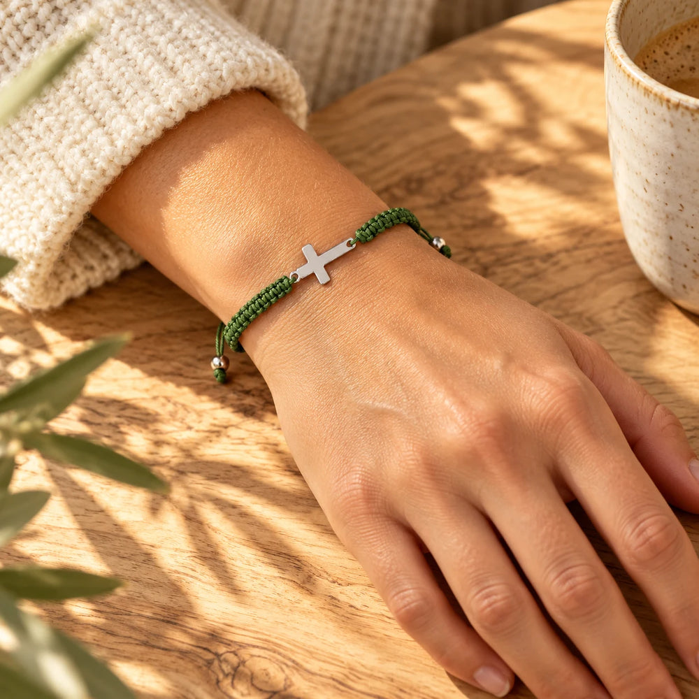 Olive-green cord cross bracelet on a wrist beside a ceramic cup — true size: the braided cord is about 5 mm thick and the sideways steel cross about 20 × 11 mm